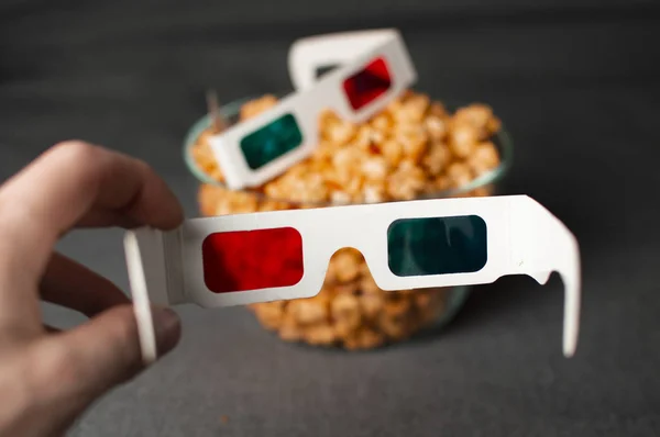 Gafas 3D y palomitas de maíz cursi se encuentran en un plato de vidrio en una cama gris, una persona se pone gafas —  Fotos de Stock