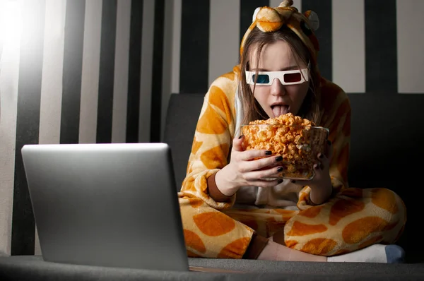 Genç kız pijamalarıyla evde film izliyor, 3D gözlük takıyor, patlamış mısır yiyor ve kanepede oturuyor. — Stok fotoğraf