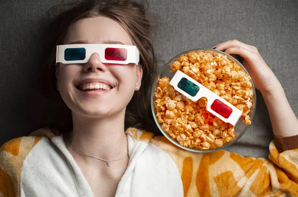 Young cute girl lies on a gray background in 3d glasses eats popcorn and watches a movie — 스톡 사진