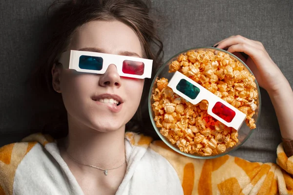 Young cute girl lies on a gray background in 3d glasses eats popcorn and watches a movie — 스톡 사진