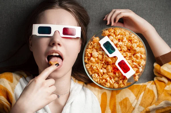 Young cute girl lies on a gray background in 3d glasses eats popcorn and watches a movie — 스톡 사진