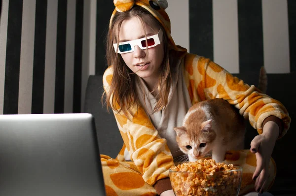 Genç kız pijamalarıyla evde film izliyor, 3D gözlük takıyor, patlamış mısır yiyor ve kanepede oturuyor.