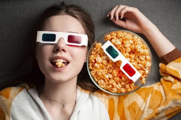 Young cute girl lies on a gray background in 3d glasses eats popcorn and watches a movie — 스톡 사진