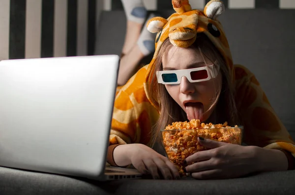 Молодая девушка смотрит кино дома в пижаме в 3D очках, ест попкорн и сидит на диване в комнате — стоковое фото