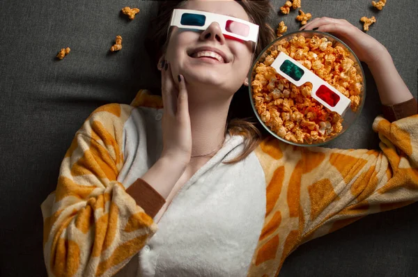 Young beautiful alone girl in 3d glasses is laying on a gray background, eating cheesy popcorn and watching a movie at home on her bed — 스톡 사진