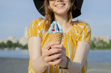 Sahilde sarı çizgili tişörtlü, turuncu gözlüklü, siyah şapkalı ve dondurma yiyen genç ve güzel bir kız yazın şehir dışında yürüyor.