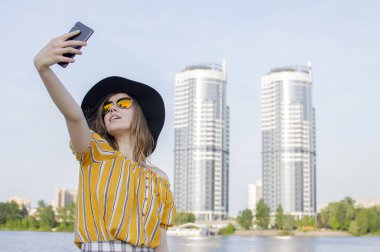 Şapkalı ve gözlüklü genç bir kız şehrin arka planında selfie çekiyor. Seyahat ediyor ve yürüyor.