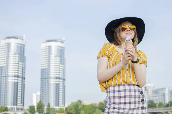 Young beautiful girl standing on the beach in a yellow striped T-shirt in orange glasses and a black hat and eating ice cream, walking in the summer outside the city — 스톡 사진