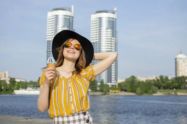 Молодая красивая девушка стоит на пляже в желтой полосатой футболке в оранжевых очках и черной шляпе и ест мороженое, гуляя летом за городом — стоковое фото