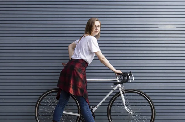 Όμορφο νεαρό κορίτσι στέκεται με ένα λευκό ποδήλατο σε φόντο γκρίζο ριγέ τοίχο, η γυναίκα είναι ευτυχισμένη — Φωτογραφία Αρχείου