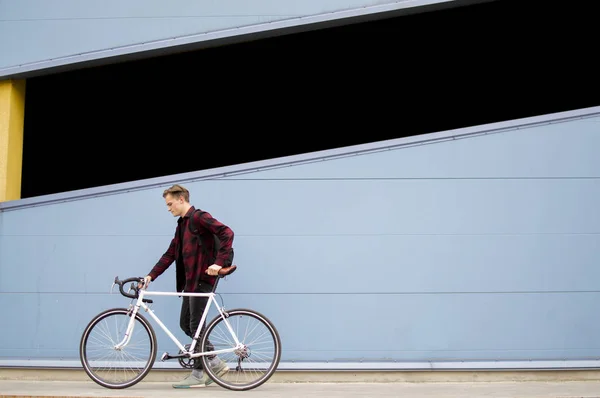 Young stylish guy walks with a white bicycle in front of a blue wall, a student with a backpack returns — Stock Photo, Image