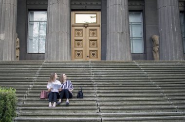 İki güzel öğrenci okulun yakınındaki merdivenlerde oturup ellerinde kağıt tutar ve ders öğrenirler, arada üniversitede iletişim kurarlar.