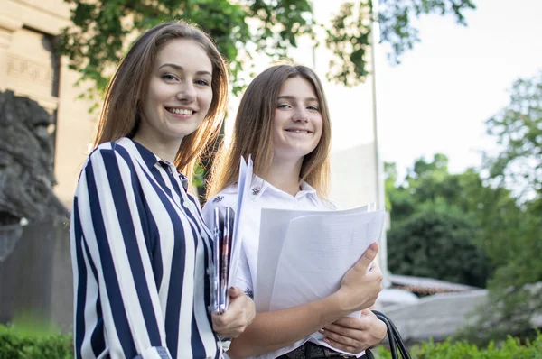 两个漂亮的学生站在大学附近，手里拿着论文，对着大学的背景微笑着，他们去上学 — 图库照片