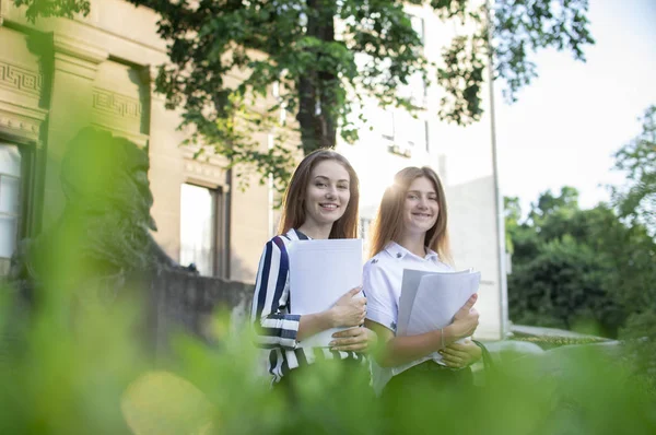 两个漂亮的学生站在大学附近，手里拿着论文，对着大学的背景微笑着，他们去上学 — 图库照片