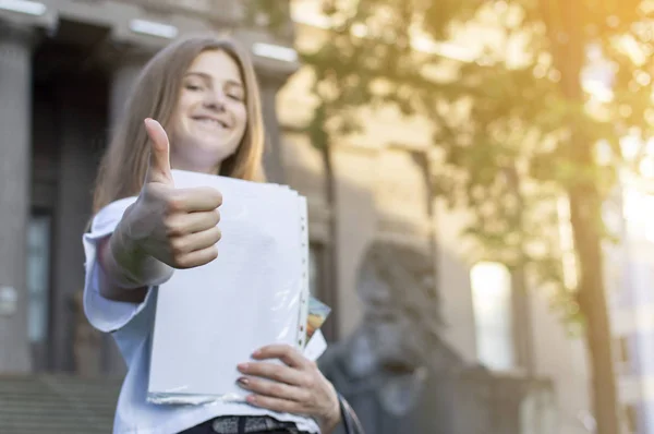 漂亮的学生站在大学附近，手里拿着一张纸微笑着，像在大学的背景，她去学校 — 图库照片