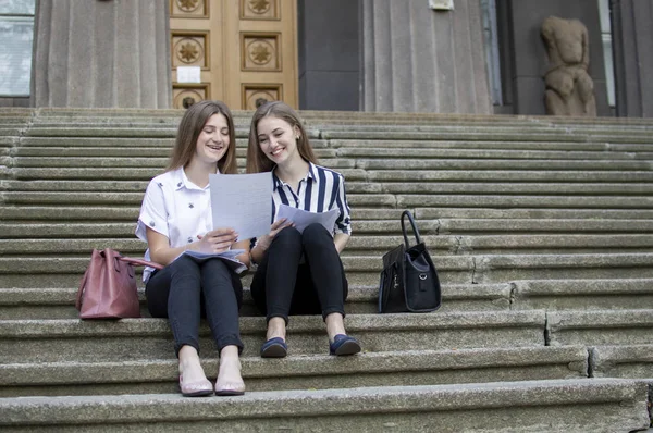 Δύο όμορφοι μαθητές κάθονται στα σκαλιά κοντά στο σχολείο, κρατούν χαρτί στα χέρια τους και μαθαίνουν μαθήματα, επικοινωνούν στο Πανεπιστήμιο κατά τη διάρκεια του διαλείμματος — Φωτογραφία Αρχείου