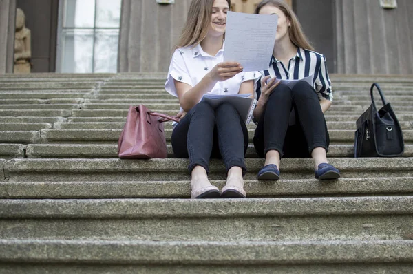 Δύο όμορφοι μαθητές κάθονται στα σκαλιά κοντά στο σχολείο, κρατούν χαρτί στα χέρια τους και μαθαίνουν μαθήματα, επικοινωνούν στο Πανεπιστήμιο κατά τη διάρκεια του διαλείμματος — Φωτογραφία Αρχείου