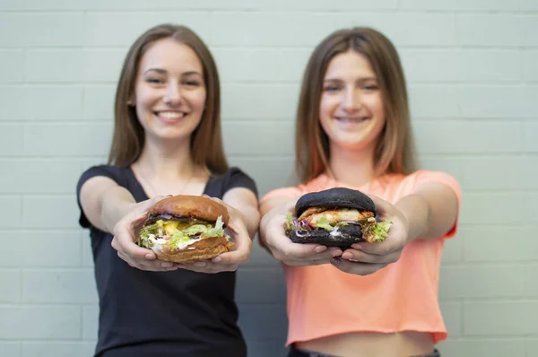 젊은 아름다운 여자 - 학생들은 푸른 벽 근처에 서서 햄버거와 미소를 잡고, 그들은 행복하고 카메라에 치즈 버거를 보여줍니다 — 스톡 사진