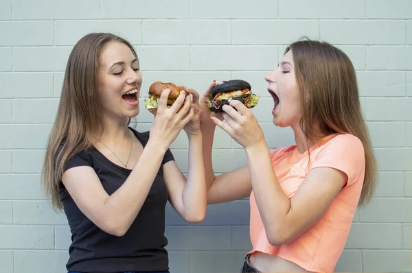 Молоді красиві дівчата-студенти стоять біля синьої стіни, тримають бургерів і посміхаються, вони їдять великих сирників, відкриваючи рот широким — стокове фото