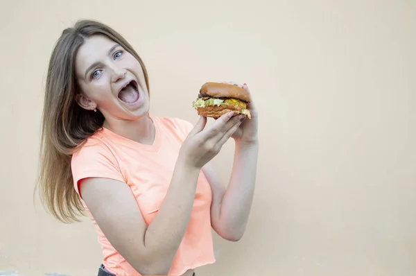 attractive teen girl stands against the orange wall in an orange T-shirt and holds a big burger in her hand, she screams and opens her mouth wide