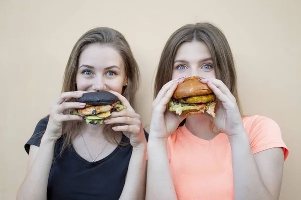 젊고 아름다운 여학생들 이 오렌지 벽 근처에 서서 햄버거를 들고 미소짓습니다. 패스트푸드를 먹으면 행복해 합니다. — 스톡 사진