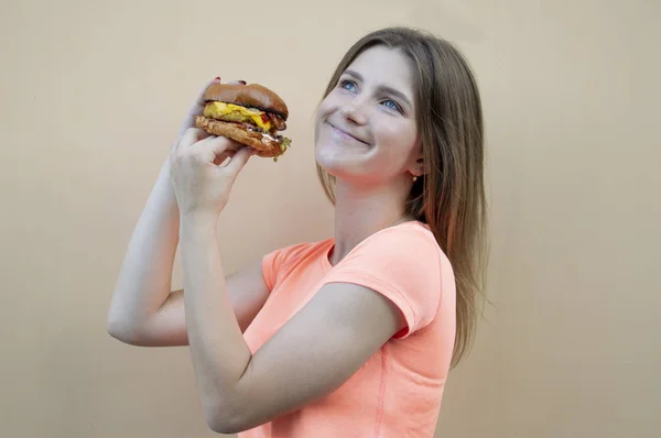 Приваблива дівчина-підліток стоїть на тлі помаранчевої стіни в помаранчевій футболці і тримає великий бургер в руці, вона посміхається і пахне чізбургер — стокове фото