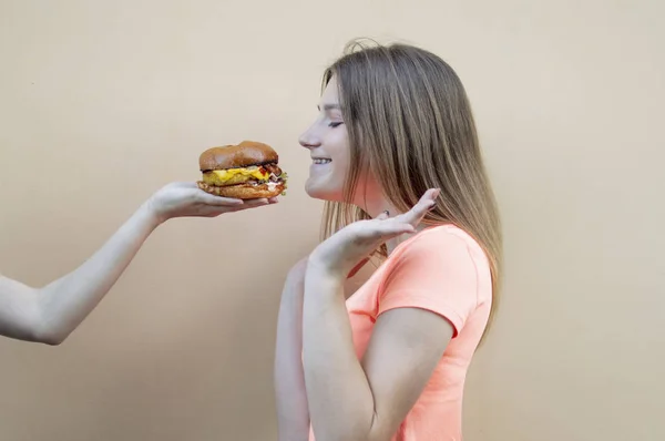 Приваблива дівчина-підліток стоїть навпроти помаранчевої стіни в помаранчевій футболці і нюхає великий бургер, інша рука дає чізбургер — стокове фото