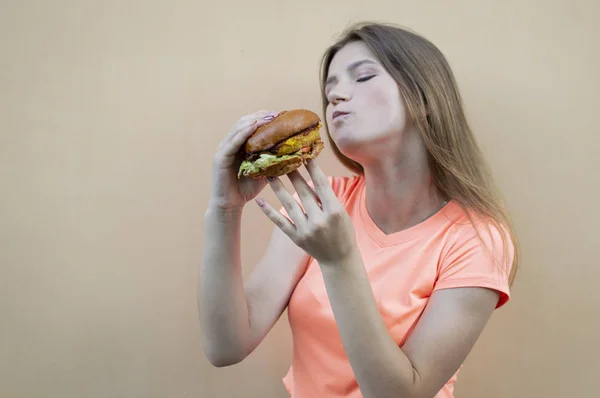 매력적인 십대 소녀는 오렌지 색 티셔츠를 입고 오렌지 벽에 서서 큰 햄버거를 손에 들고 입을 크게 벌리고 먹습니다. — 스톡 사진