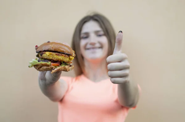 attractive teen girl stands against an orange wall in an orange T-shirt and holds a big burger, she shows a like with her hand