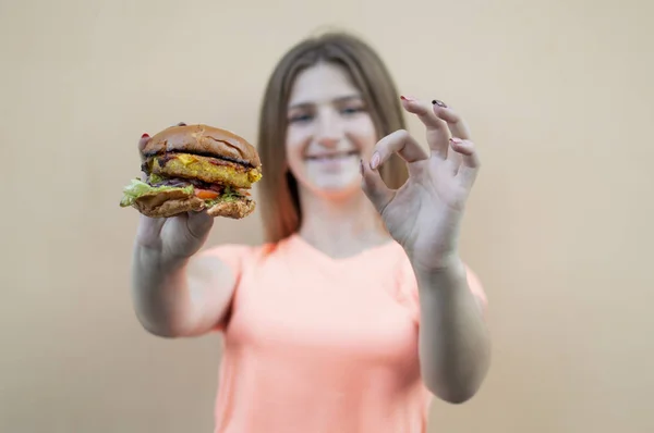 Attraktive Teenie-Mädchen steht vor dem Hintergrund einer orangefarbenen Wand in einem orangefarbenen T-Shirt und hält einen großen Burger, sie zeigt ihr Okay mit der Hand — Stockfoto