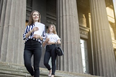 Iki güzel öğrenci, ellerinde kağıtları tutarak ve üniversitenin arka plan karşı gülümseyerek, üniversitenin yakınında duruyor, onlar okula gitmek