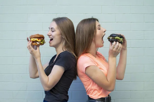 Young beautiful girls-students stand near the blue wall, hold burgers and smile, they eat big cheeseburgers, opening their mouth wide — 스톡 사진