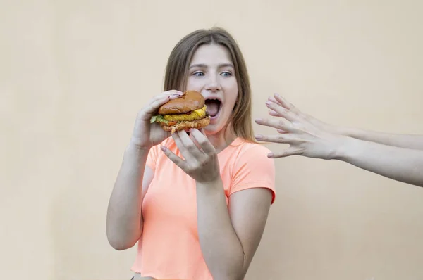 Young teen girl stands against the background of an orange wall in an orange T-shirt and holds a big burger in her hand, the other hand selects a cheeseburger — 스톡 사진