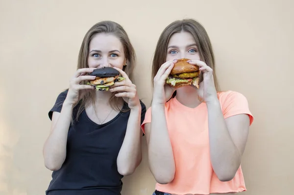 젊고 아름다운 여학생들 이 오렌지 벽 근처에 서서 햄버거를 들고 미소짓습니다. 패스트푸드를 먹으면 행복해 합니다. — 스톡 사진