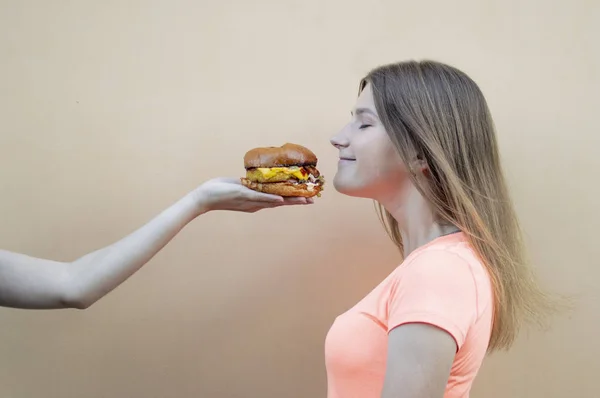매력적인 십대 소녀는 오렌지 색 티셔츠에 오렌지 벽에 서서 큰 햄버거를 냄새, 다른 한편으로는 치즈 버거를 제공합니다 — 스톡 사진