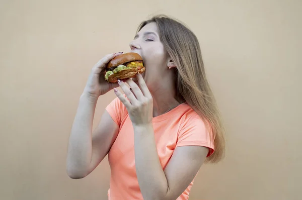 Menina adolescente atraente fica contra a parede laranja em uma camiseta laranja e segura um grande hambúrguer na mão, ela abre a boca e come — Fotografia de Stock