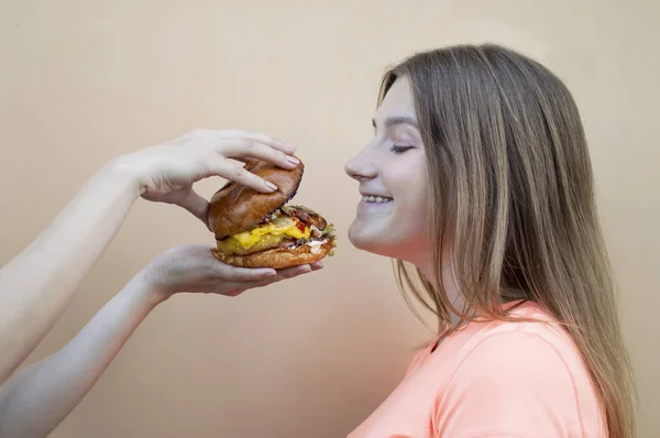 Приваблива дівчина-підліток стоїть на тлі помаранчевої стіни в помаранчевій футболці і тримає великий бургер в руці, вона посміхається і пахне чізбургер — стокове фото