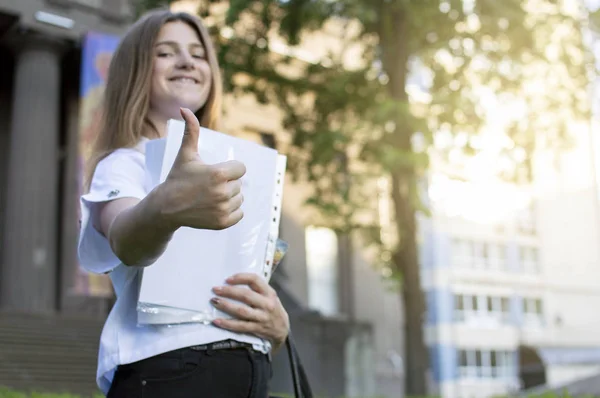 Όμορφη μαθήτρια στέκεται κοντά στο Πανεπιστήμιο, κρατώντας ένα χαρτί στα χέρια της χαμογελώντας και δείχνοντας σαν στο φόντο του κολλεγίου, πηγαίνει στο σχολείο — Φωτογραφία Αρχείου