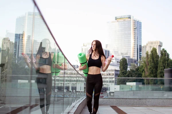 Gadis atletik melakukan yoga di pagi hari di jalan, dia memegang tikar dan botol di latar belakang kota, wanita kebugaran pergi ke gym, potret — Stok Foto