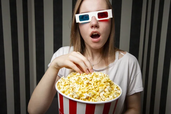 Young girl watching a movie and eating popcorn wearing 3d glasses against a striped wall at night, she is emotional and surprised — Stock Photo, Image