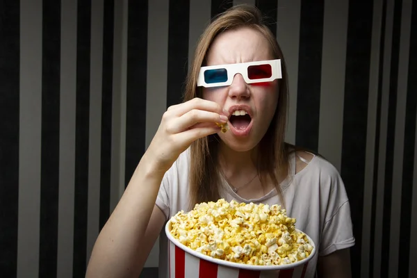 young girl watching a movie and eating popcorn wearing 3d glasses against a striped wall at night, she is emotional and surprised