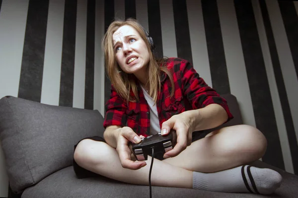 Jovem menina senta-se no sofá e joga o console no joystick, ela está focada em jogos de vídeo — Fotografia de Stock