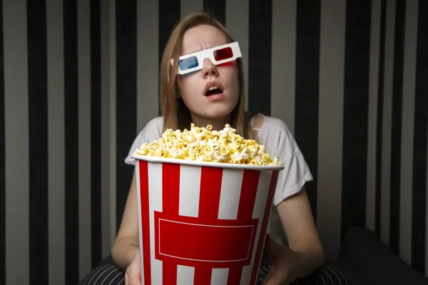 映画を見て、夜にストライプの壁に3Dメガネをかけてポップコーンを食べている若い女の子は、彼女は感情的で驚いています — ストック写真