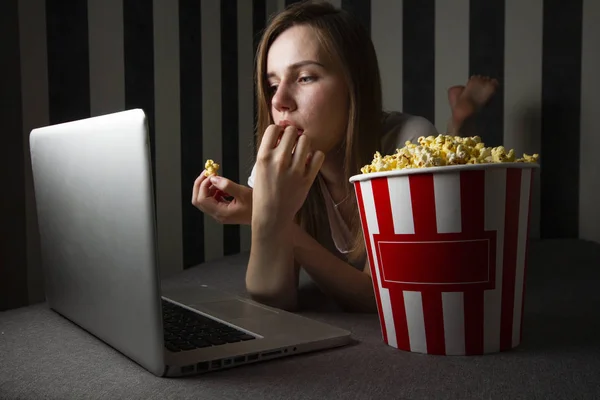 A young girl watching a TV show at night in a laptop and eating popcorn, she uses a computer while lying on the sofa — Stock Photo, Image