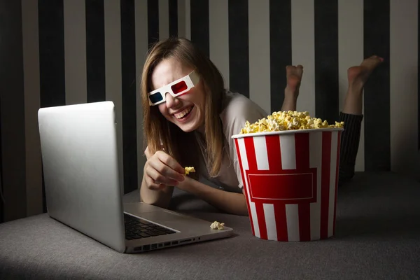 Una chica joven viendo un programa de televisión por la noche en una computadora portátil y comiendo palomitas de maíz, usa una computadora mientras está acostada en el sofá — Foto de Stock