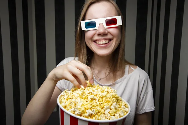 Young girl watching a movie and eating popcorn wearing 3d glasses against a striped wall at night, she is emotional and surprised — Stock Photo, Image