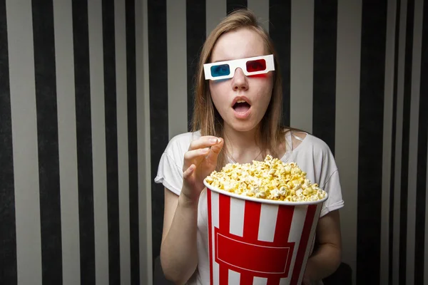 Young girl watching a movie and eating popcorn wearing 3d glasses against a striped wall at night, she is emotional and surprised — Stock Photo, Image