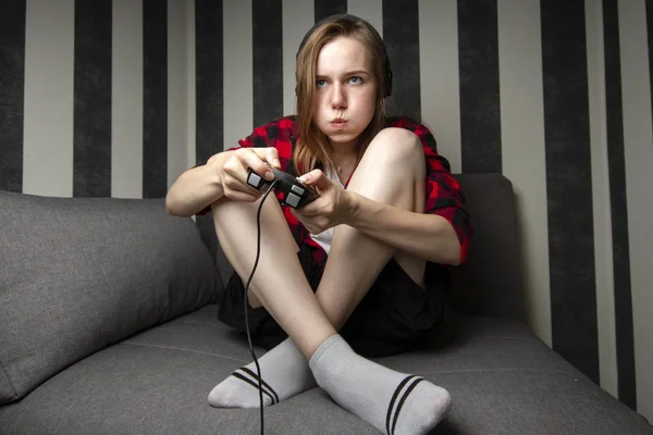 Jovem menina senta-se no sofá e joga o console no joystick, ela está focada em jogos de vídeo — Fotografia de Stock