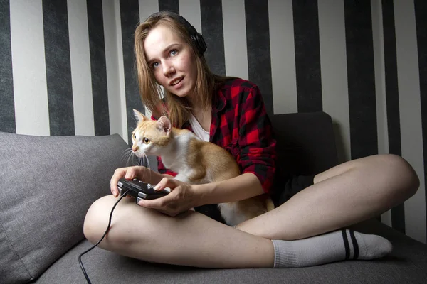 Menina alegre sentado no sofá e jogar videogames com um gato, ela segura o joystick e sorri para a câmera — Fotografia de Stock