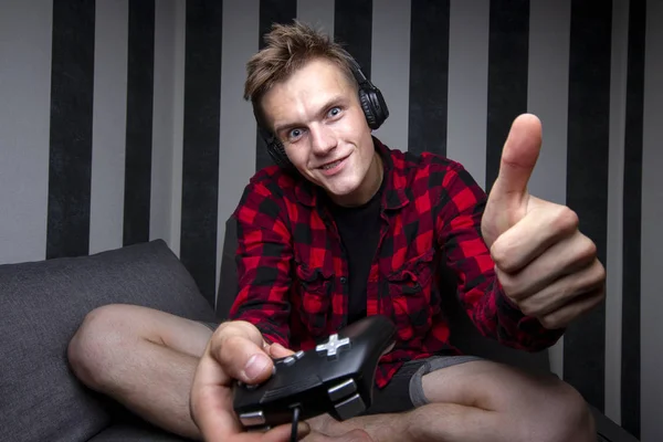 Jovem cara joga jogos de vídeo no console, ele se senta em casa no sofá e segura o joystick à noite — Fotografia de Stock